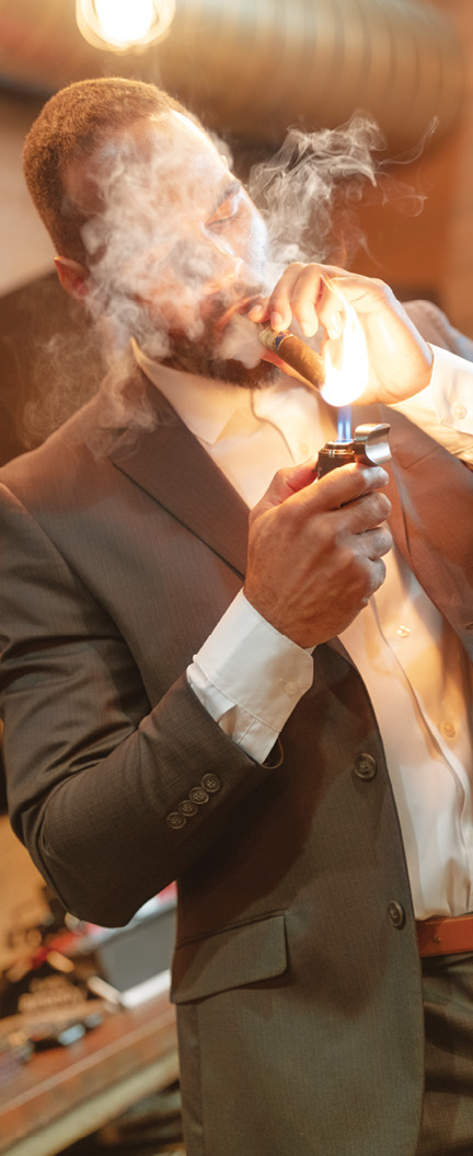 A person wearing a suit jacket and white shirt is lighting a cigar with a lighter while smoke surrounds their face.

Description generated by AI